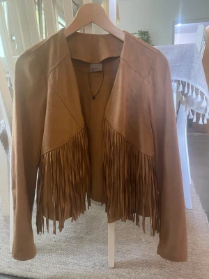 Brun jacka fransar - Snygg brun mocka jacka med långa fransar framtill. Jackan har öppen front utan dragkedja eller knappar och en avslappnad passform. Perfekt för dig som vill ha en boho-inspirerad look och gillar detaljer som sticker ut.