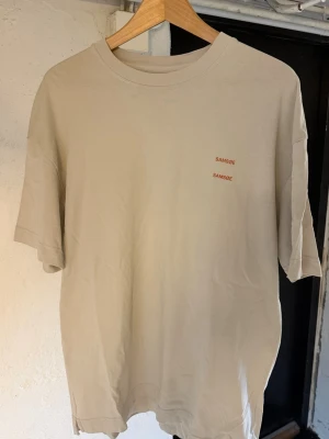 Beige t-shirt från Samsøe Samsøe - Säljer en beige t-shirt från Samsøe Samsøe med diskret logga i rött på bröstet. T-shirten har rund hals, korta ärmar och en relaxed passform. Perfekt basplagg i mjuk bomull som funkar till allt. Använd ett par gånger 