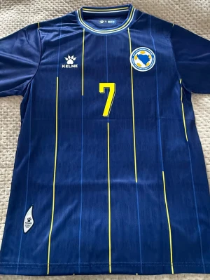 Blå fotbollströja Bosnien #7 Dedic - Snygg blå fotbollströja från Kelme med gula och blå ränder, nummer 7 och namnet Dedic på ryggen. Bröstet har Bosnien och Hercegovinas landslagsemblem. Perfekt för dig som vill stötta laget på match eller träning.