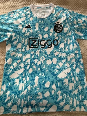 Ajax blåvit matchtröja - Säljer en Ajax matchtröja från Adidas i blått och vitt med ett unikt abstrakt mönster. Tröjan har korta ärmar, rund hals och Ajax-logga samt Ziggo-tryck på bröstet. Perfekt för dig som älskar fotboll och vill sticka ut på planen.