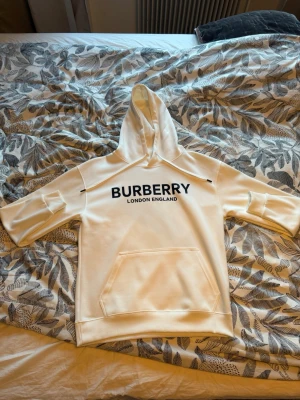 Vit Burberry hoodie med tryck - Snygg vit hoodie från Burberry med svart text 'BURBERRY LONDON ENGLAND' på bröstet. Hoodien har huva med dragsko, korta ärmar och en stor magficka. Perfekt för dig som vill ha en stilren och trendig look.