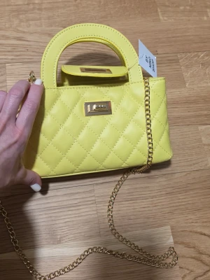 Gul quiltad handväska med guldkedja - Trendig gul handväska i quiltat konstläder med guldfärgad kedjerem och vridlås. Väskan har en kompakt, rektangulär form och ett kort handtag upptill. Perfekt för dig som vill sticka ut med en färgglad accessoar.