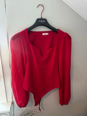 Röd blus med v-ringning från Nelly - Snygg röd blus från Nelly med långärmade, lätt transparenta ärmar och markerad v-ringning. Toppen har en asymmetrisk nederkant och är tillverkad i ett tunt, flowy material som ger en trendig look.
