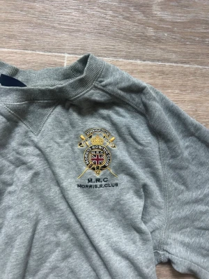 Grå sweatshirt från Morris R. Club - Snygg grå sweatshirt från Morris R. Club med broderad logga och brittisk flagga på bröstet. Tröjan har rund halsringning och ribbade muddar. Perfekt för en avslappnad och sportig stil.