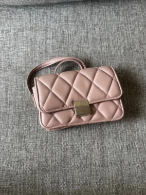 Rosa quiltad handväska med axelrem - Snygg rosa handväska i quiltad design med rutmönster och silverfärgat spänne framtill. Väskan har en smidig axelrem och kompakt, rektangulär form. Perfekt för dig som gillar stilrena accessoarer i mjukt material.