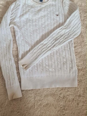Vit stickad tröja - Kabelstickad vit tröja med rund halsringning och ribbade muddar. Tröjan har ett diskret broderat märke på bröstet. Fläck på insidan