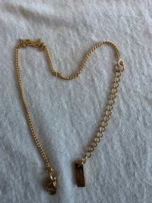 Guldigt halsband från Yves Saint Laurent - Snyggt guldigt halsband från Yves Saint Laurent med tunn kedja och YSL-logga som detalj. Halsbandet har justerbar längd och en liten bricka med ingraverad text. Perfekt för dig som gillar stilrena accessoarer med lyxig känsla.