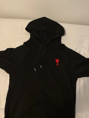 Svart hoodie från Ami med rött hjärta - Säljer en svart hoodie från Ami med den ikoniska röda hjärtemärket broderat på bröstet. Hoodien har huva med dragsko och en stor magficka. Perfekt för dig som gillar stilrena plagg med en twist. Materialet känns mjukt och skönt.