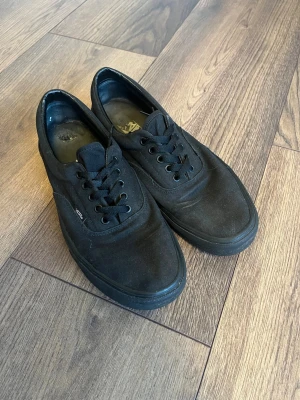 Svarta Vans sneakers i canvas - Klassiska svarta Vans sneakers i canvas med lågt skaft och svarta skosnören. Skorna har en enkel och stilren design med Vans-logga på sidan och röd detalj på hälen. Perfekta för en avslappnad streetwear-look.