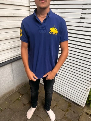 Vintage Ralph Lauren pike - Snygg marinblå pikétröja med stor gul logga på bröstet och siffran 3 i gult på ärmen. Klassisk krage och kortärmad modell, perfekt till jeans eller shorts. Tillverkad i mjuk bomull för skön känsla.