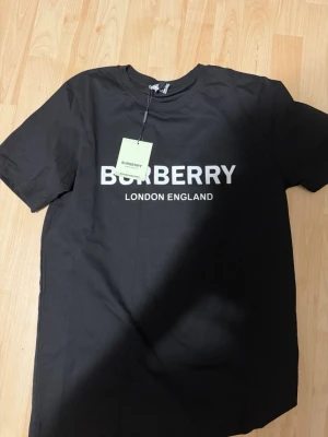 Svart Burberry t-shirt med logga - Svart t-shirt från Burberry med klassisk vit logga 'BURBERRY LONDON ENGLAND' tryckt på bröstet. T-shirten har rund halsringning och korta ärmar. Perfekt för dig som vill ha en stilren och trendig look. Äkta 