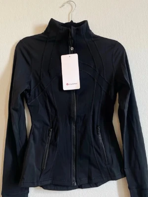 Svart träningsjacka från Lululemon st 4 - Snygg svart träningsjacka från Lululemon med figurnära passform, hög krage och dragkedja framtill. Jackan har långa ärmar och två fickor med dragkedja. Perfekt för dig som vill ha en sportig och stilren look.