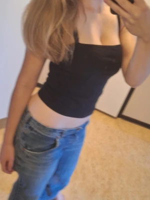 Svart croppad linne med smala axelband - Snyggt svart croppat linne med smala axelband och rak halsringning. Perfekt till jeans eller kjol för en enkel och stilren look. Mjukt och stretchigt material som sitter bekvämt på kroppen. Detaljerna på axelbanden på ryggen är en del av min bh och inte linnet så ni vet🫶🏻