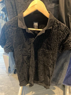 Svart polotopp från Black Squad XS - Säljer en svart polotopp från Black Squad i storlek XS. Toppen har ett unikt strukturerat mönster i tyget och klassisk krage med knappar framtill. Slim fit-modell som sitter snyggt på kroppen och passar perfekt till jeans eller kjol.
