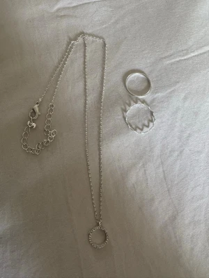 Gulligt Smyckeset med halsband och ringar - Säljer ett stilrent smyckeset i silverfärg som består av ett halsband med cirkelhänge och två matchande ringar!🤍Halsbandet har en tunn kedja och cirkeln är dekorerad med små stenar/diamanter. Ringarna är enkla, en slät och en med vågig design. Perfekt för dig som gillar minimalistiska accessoarer😍Smyckena passar bra till allting, uppklädd eller nerklädd. Så fina! Tror allt är från H&M. Hör av vid funderingar eller bud, rabbat på bundles❤️Säljer mer smycken på min profil. 