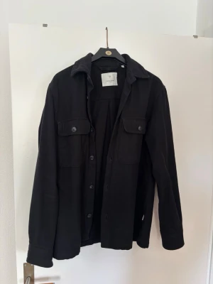 Svart overshirt från Jack & Jones - Säljer en svart overshirt från Jack & Jones med klassisk krage, knäppning framtill och två bröstfickor med lock och knapp. Overshirten har långa ärmar och är tillverkad i ett mjukt tyg som passar perfekt för lager på lager.