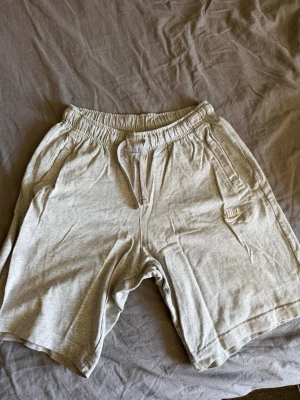 Grå shorts från Nike med snörning - Snygga grå shorts från Nike i mjuk bomull med elastisk midja och snörning. Shortsen har fickor på sidorna och en liten Nike-logga på vänster ben. Perfekta för chill dagar eller träning.
