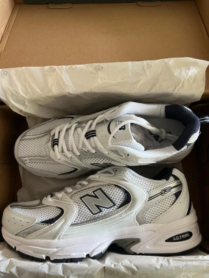 New Balance 530 vita sneakers - Snygga New Balance 530 sneakers i vitt med silver- och marinblå detaljer. Ovandelen är i mesh och syntetmaterial för en sportig look. Klassisk N-logga på sidan och ABZORB-dämpning i sulan för extra komfort. Perfekta för en trendig och avslappnad stil.