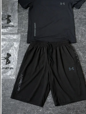 Svart träningsset från Under Armour - Säljer ett svart träningsset från Under Armour med t-shirt och shorts. Båda plaggen har logga och tryck i grått. Materialet är lätt och andas, perfekt för träning eller gym. Shortsen har resår och dragsko i midjan för skön passform.