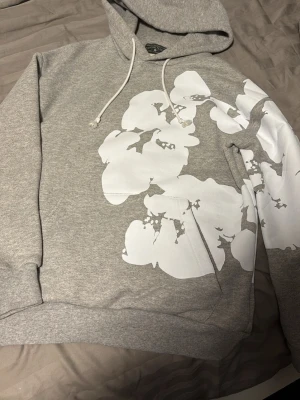 Grå hoodie med blommor från Denim Tears - Snygg grå hoodie från Denim Tears med stora vita blommor tryckta över framsidan och ena ärmen. Klassisk huva med dragsko och mjukt innerfoder. Perfekt för dig som vill sticka ut med en unik och trendig look.
