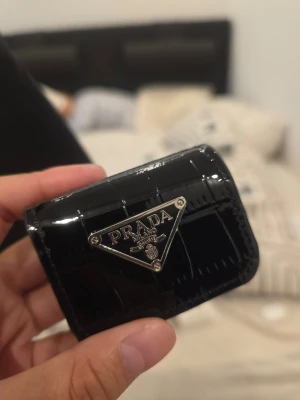 Svart AirPods-fodral PD - Lyxig svart Airpods fodral som passar alla modeller från PD i glansigt krokopräglat läder. Framsidan har en ikonisk silverfärgad metallogga och baksidan en silverkedja med karbinhake, perfekt att fästa på väska eller nyckelknippa. Kompakt och stilren design.