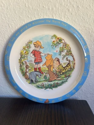 Nalle puh tallrik  - Nalle Puh tallrik med motiv av Nalle Puh och hans vänner. Tallriken är vit med en blå kant och texten "We are going to discover the North Pole" explained Piglet.  Obs! Skick kan ses på bilder! Inga större skavanker.