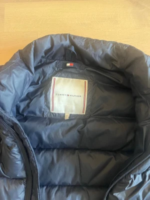 Mörkblå dunväst från Tommy Hilfiger stl 152 - Snygg mörkblå dunväst från Tommy Hilfiger med quiltad design och hög krage. Diskret logga på insidan och Hilfiger-tryck längs ärmöppningarna. Perfekt för lager på lager och streetwear-stil.