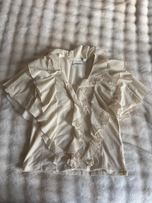Krämvit volangblus från Kappahl - Supersöt krämvit blus från Kappahl i storlek XS. Blusen har korta ärmar och är dekorerad med stora volanger längs halsringningen och framtill. Tillverkad i ett tunt, lätt tyg som ger en romantisk och luftig känsla.