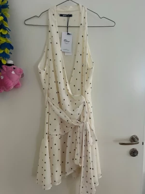 Vit prickig klänning - Supersöt vit kortklänning med svarta prickar och omlottdesign. Klänningen har djup v-ringning, knyt i midjan och volangkant nedtill. Ärmlös modell med lätt och luftig känsla, perfekt för varma dagar. Helt nytt, oanvänt klänning.  Storlek S. 