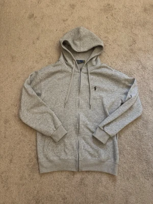 Grå zip hoodie från Polo Ralph Lauren - Snygg grå hoodie med dragkedja från Polo Ralph Lauren. Klassisk design med huva och dragsnören, ribbade muddar och fickor framtill. Ikoniska svarta loggan broderad på bröstet. Perfekt till chill eller streetwear-stil.