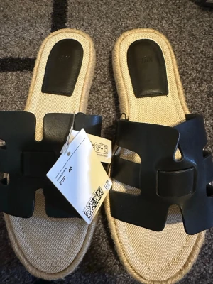 Svarta sandaler från H&M, storlek 40 - Snygga sandaler från H&M i storlek 40 med svart bred rem i syntetmaterial och naturfärgad sula med flätad kant. Perfekt för sommarens alla äventyr, enkel slip-on modell med platt sula och stilren design.