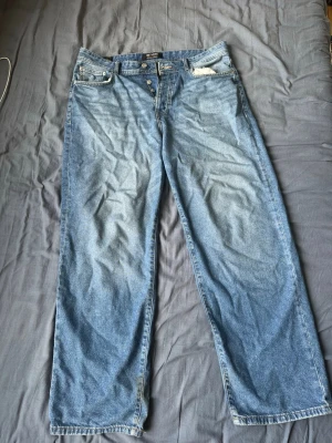 Blåa raka jeans med knappgylf Only&Sons - Säljer ett par blåa jeans med rak passform och knappgylf. Jeansen har klassisk femficksdesign och ljus tvätt med slitningar vid fickorna. Materialet är denim i bomull och benen är vida hela vägen ner.