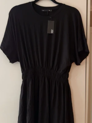 Svart kortärmad klänning  - En svart klänning med rund halsringning och korta ärmar. Klänningen har resår i midjan som ger en snygg siluett och är tillverkad i ett mjukt tyg. Enkel och stilren design som passar till många tillfällen.