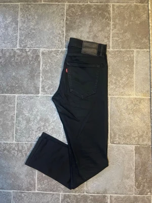 Svarta slim fit jeans - Snygga svarta slim fit jeans med klassisk femficksdesign och diskret röd detalj vid bakfickan. Jeansen har raka ben och är tillverkade i ett mjukt denimtyg som ger en skön passform. Perfekta för dig som gillar en stilren och enkel look.