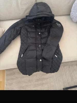 Svart dunjacka med huva - Snygg svart dunjacka med huva från RockAndBlue och quiltade detaljer på sidorna och ärmarna. Jackan har dragkedjor på fickorna, knappar framtill och en patch på ena ärmen. Perfekt för kalla vinterdagar och har en figurnära passform. Finns ett litet håll men går att sy ihop