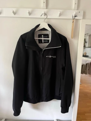 Svart jacka från Sail Racing - Stilren svart jacka från Sail Racing med dragkedja framtill och hög krage. Jackan har logga på bröstet och ribbade muddar vid ärmslut och nederkant. Perfekt för dig som gillar en sportig och enkel look.