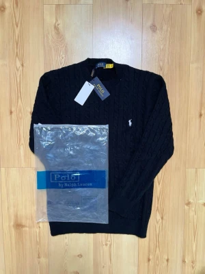 Svart kabelstickad tröja Polo Ralph Lauren - Kabelstickad svart tröja från Polo Ralph Lauren med rund halsringning och den klassiska broderade logotypen på bröstet. Tröjan har långa ärmar och ribbade muddar vid ärmslut och nederkant. Perfekt för dig som gillar stilren och tidlös design.