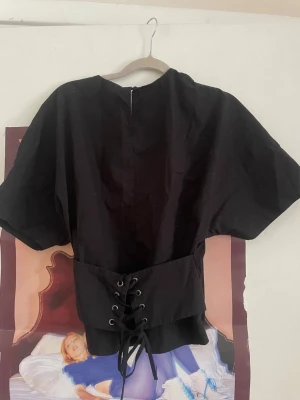 Svart topp med snörning från GAP - Snygg svart topp från GAP med korta ärmar och cool snörningsdetalj i midjan. Toppen har en rak passform och dragkedja i nacken. Perfekt för dig som gillar en edgy och modern stil.