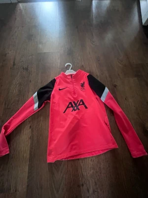 Röd Nike Liverpool träningströja - Snygg röd långärmad träningströja från Nike med Liverpool FC-logga och svart AXA-tryck på bröstet. Tröjan har svarta och vita detaljer på ärmarna samt halv dragkedja framtill. Tillverkad i Dri-FIT-material som andas och passar perfekt till träning.