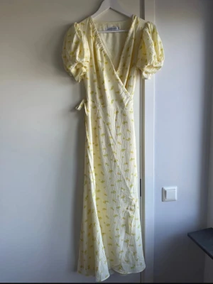 Ljusgul omlottklänning från Adoore - Supersöt ljusgul långklänning från Adoore med puffärmar, omlottmodell och knyt i midjan. Klänningen har ett diskret blommigt mönster och v-ringad hals. Perfekt för sommarens alla tillfällen! Använd 1 gång!