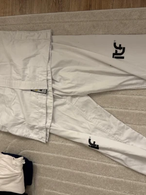 Vit Taekwondo-dräkt ITF - Säljer en klassisk vit Taekwondo-dräkt med svarta ITF-tryck på byxbenen och stor Taekwon-Do ITF-logga på ryggen. Dräkten är tillverkad i slitstarkt bomullstyg och har traditionell passform med knytband i midjan. Perfekt för träning eller tävling.