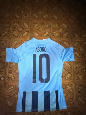 Djurgården matchtröja Asoro #10 - Säljer en ljusblå Djurgården fotbollströja från Adidas med mörkblå ränder och klubbmärke på bröstet. Tröjan har Asoro och nummer 10 tryckt på ryggen. Perfekt för dig som vill stötta laget på match eller träning.