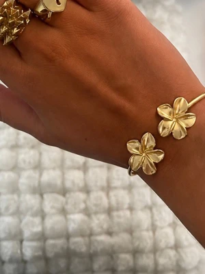 Guldigt armband med blommor - Snyggt armband i guldfärgad rostfritt metall med två stora blommor som detaljer. Blommorna har en stilren och modern design som ger armbandet en unik look. Perfekt accessoar för dig som vill sticka ut lite extra med dina smycken.