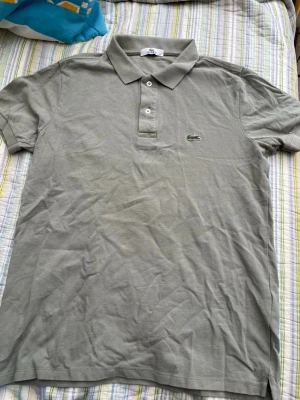 Grå pikétröja från Lacoste - Klassisk grå pikétröja från Lacoste med vit knappslå och den ikoniska gröna krokodilloggan på bröstet. Tröjan har korta ärmar och krage, tillverkad i bomull för en skön och luftig känsla.