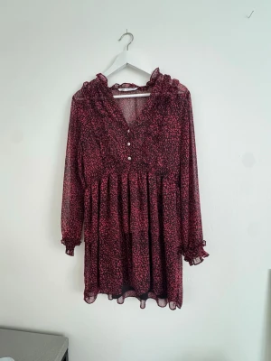 Blommig vinröd kortklänning - Söt vinröd kortklänning med små blommor, v-ringad med volangkant och knappar framtill. Klänningen har långa, transparenta ärmar med volangdetaljer vid ärmslut och en luftig passform. Perfekt för vår och sommar.