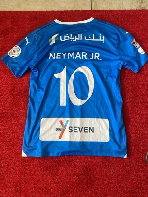Blå Al Hilal Neymar Jr. matchtröja - Snygg blå matchtröja från Al Hilal med Neymar Jr. och nummer 10 på ryggen. Tröjan har vita detaljer, flera sponsorloggor och är tillverkad i ett lätt funktionsmaterial. Perfekt för dig som älskar fotboll och vill bära din favoritspelares tröja.