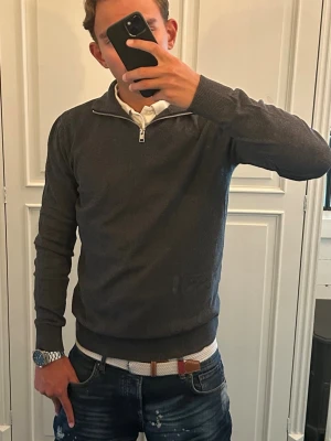 stickad halvzip tröja J&J - Snygg mörkgrå stickad tröja från Jack&Jones med halvzip och hög krage. Tröjan har ribbade muddar vid ärmslut och nederkant, perfekt för lager på lager. Passar till både skjorta och t-shirt för en clean look.