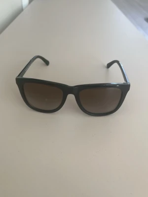 Svarta solglasögon Michael Kors - Snygga solglasögon från Michael Kors med svart båge och bruna tonade glas. Klassisk fyrkantig form och diskret logga på sidan. Perfekta för soliga dagar och ger en cool look.