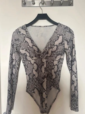 Ormmönstrad långärmad body - Snygg body med ormmönster i grått och svart. Djup v-ringning framtill och långa ärmar. Stretchigt material som sitter tajt mot kroppen och ger en cool look. Perfekt att styla med jeans eller kjol för en trendig outfit.