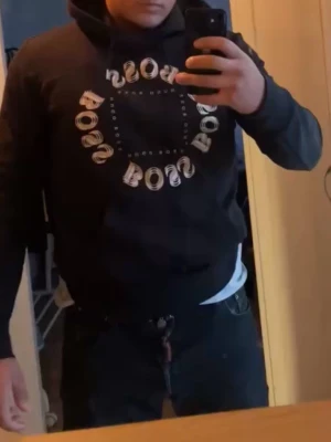 Svart hoodie med BOSS-tryck - Snygg svart hoodie från BOSS med stort cirkelformat BOSS-tryck i vitt på bröstet. Klassisk huva med dragsko och avslappnad passform. Perfekt för en casual och trendig look. Medium men kan passa en Large också jag är 85kg 174 lång.